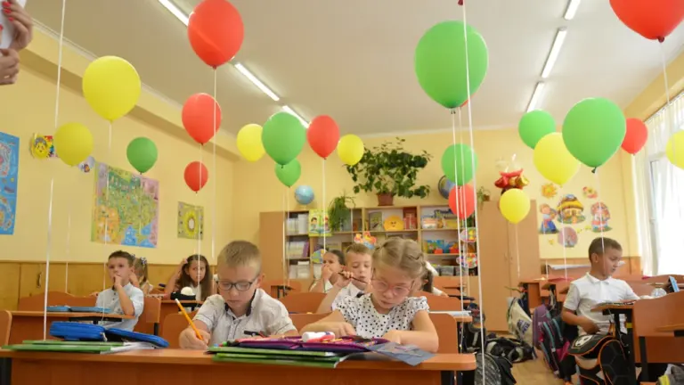 Uczniowie przy ławkach w klasie udekorowanej kolorowymi balonami, przygotowani do nauki szkolnej.